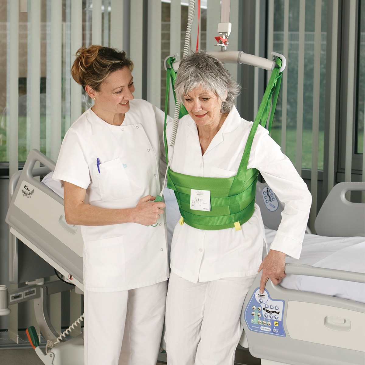 Un clinico aiuta un paziente a deambulare in una stanza d'ospedale con un sollevatore a soffitto Hillrom e MasterVest